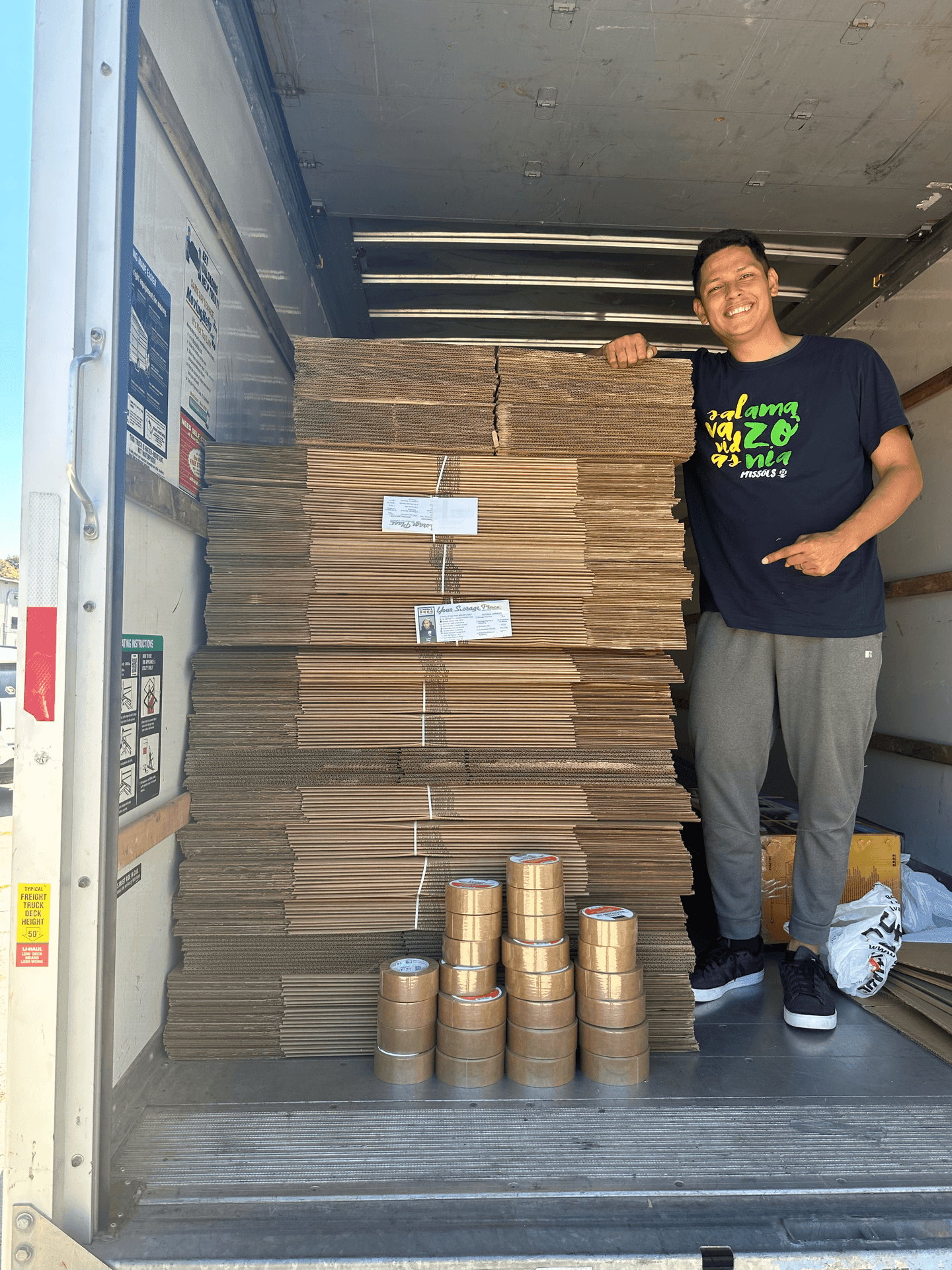 Person smiling in a truck with stacked cardboard boxes and rolls of tape.
